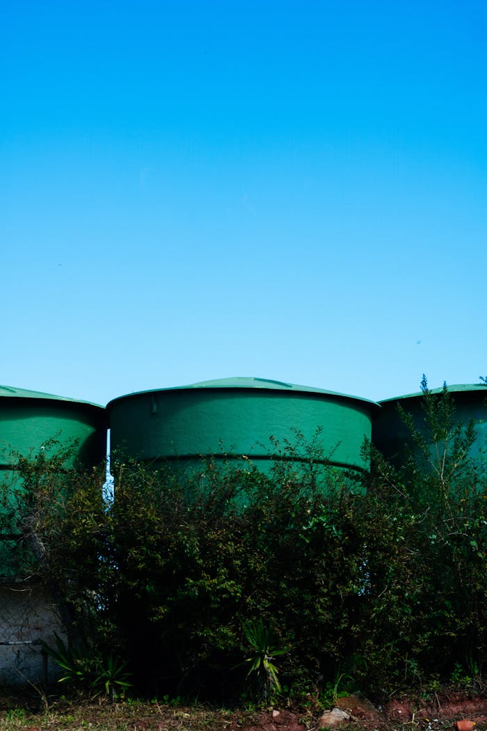 Green cylindrical water tanks in a rural area under a clear blue sky, surrounded by bushes.