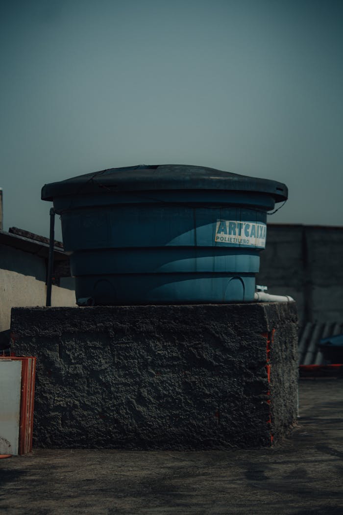 A large blue, rusty water tank positioned on a rooftop in an urban setting under a clear sky.