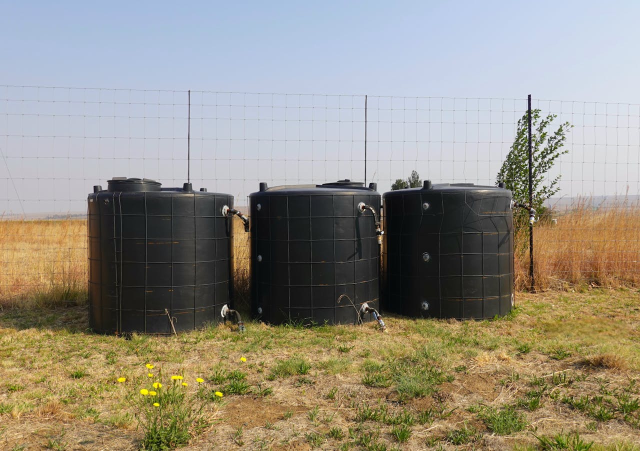 Three black water tanks in a dry, grassy field with a wire fence.